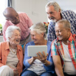 Fünf Senior*innen schauen auf den Bildschirm eines Tablets und lachen