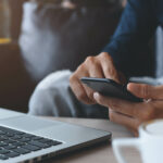 Eine Person sitzt mit dem Smartphone in der Hand vor einem aufgeklappten Laptop.